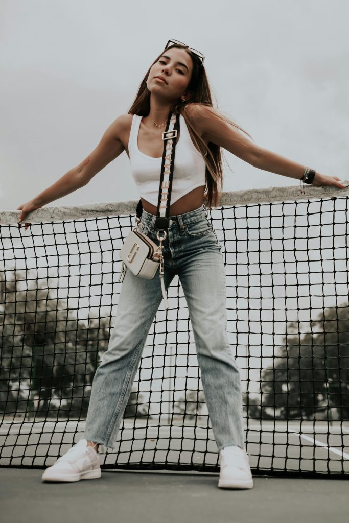 Young woman in casual wear and white sneakers posing on a tennis court, showcasing a chic and relaxed style.