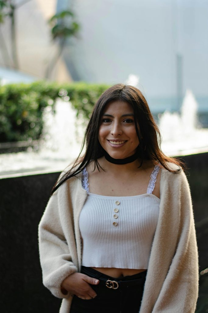 Young woman wearing a white top and beige coat, smiling outdoors near a fountain.