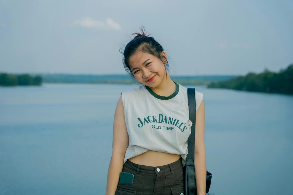 Asian woman in casual clothing posing and smiling by a lake on a sunny day.