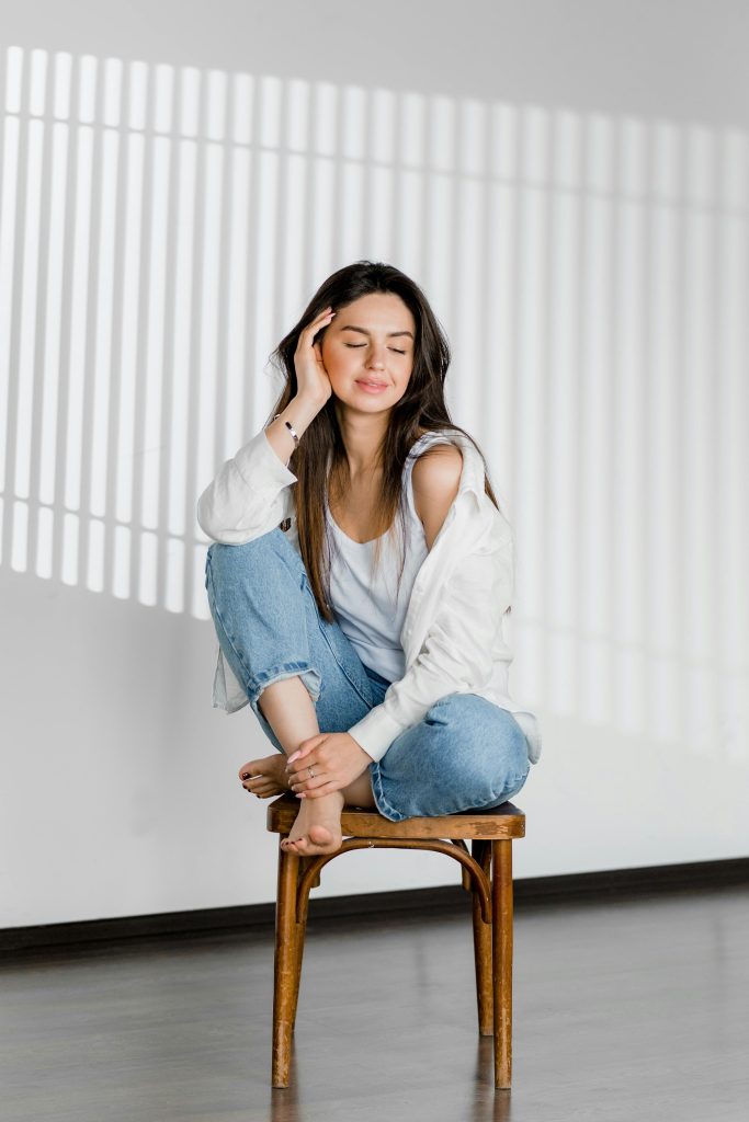A serene young woman sits barefoot on a chair, eyes closed, in a bright indoor setting.