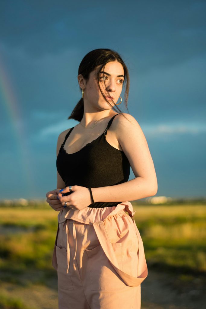 A young woman stands outside in summer light, looking away, with a rainbow in the background.