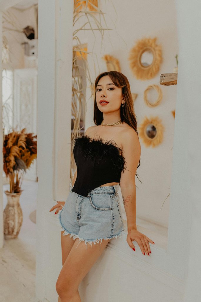 Fashionable young woman in a corset top and denim shorts posing indoors with decor.