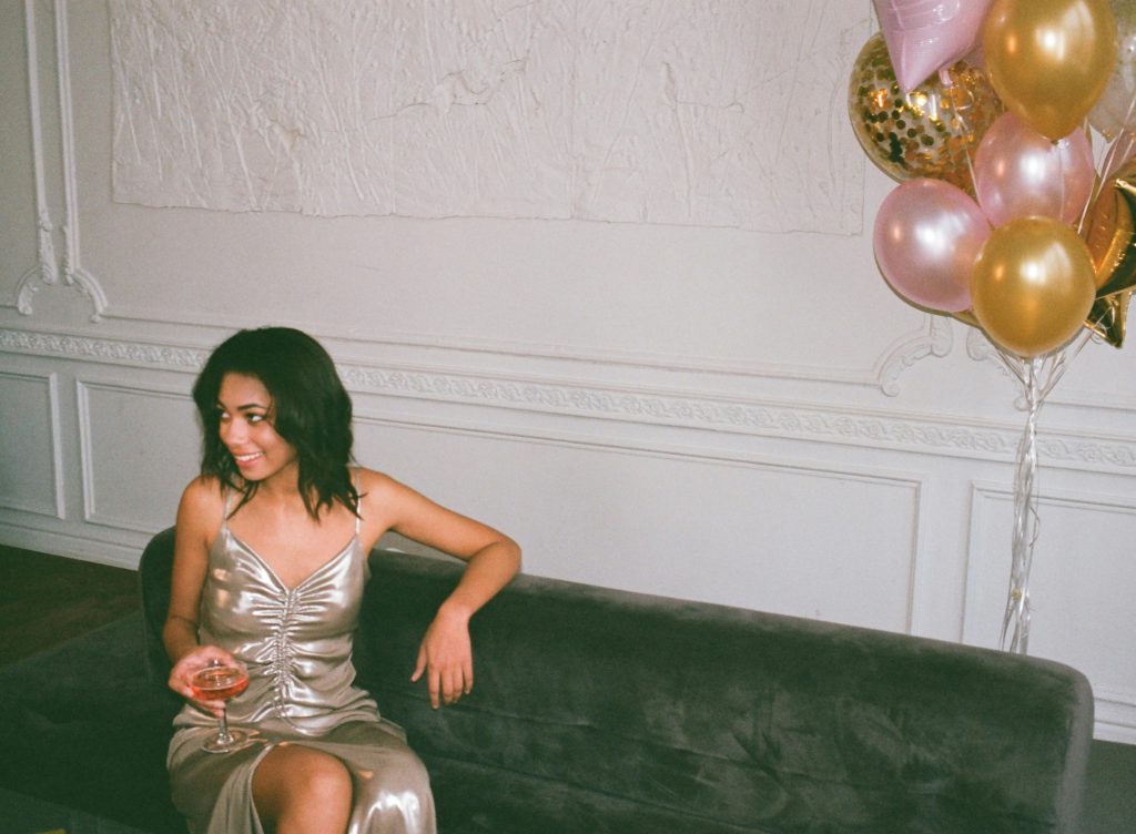 Chic woman in metallic dress sitting on sofa enjoying drinks at a party with balloons.
