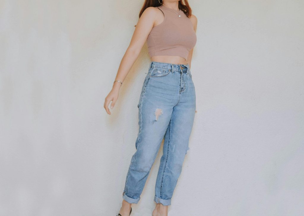 Adult in casual wear with crop top and ripped jeans, standing indoors against a neutral wall.
